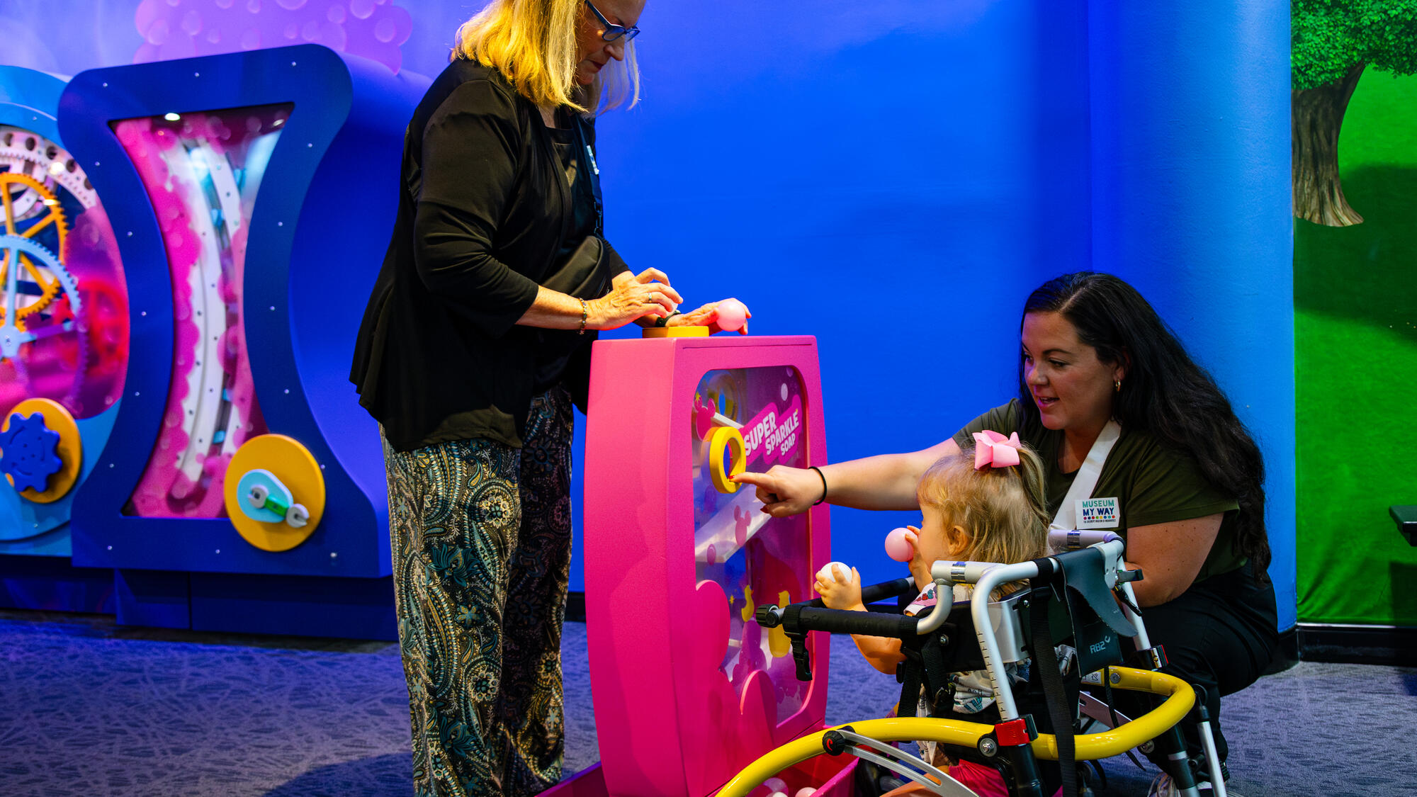 Two adults and a child using a walker play with a toddler-size interactive in the Mickey Mouse Clubhouse exhibit. The child holds two balls while one adult points where to put the balls so they can go down a track.